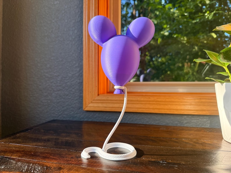 May include: A purple 3D-printed balloon sculpture featuring a rounded head with two circular ears, attached to a white, coiled, rope-like base that extends upward to support the balloon from its bottom knot, creating a floating effect.