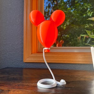 May include: A bright orange, three-dimensional balloon sculpture featuring a central rounded shape with two smaller circular ears attached at the top. A white, coiled rope extends from the base of the balloon to a circular white base.