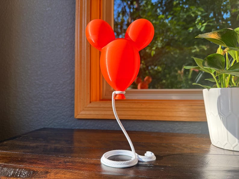 May include: A bright orange, three-dimensional balloon sculpture featuring a central rounded shape with two smaller circular ears attached at the top. A white, coiled rope extends from the base of the balloon to a circular white base.