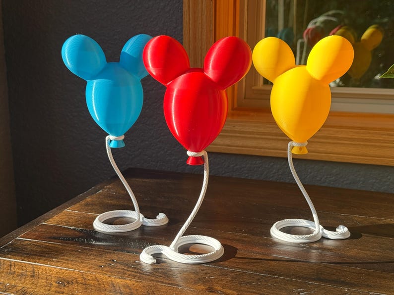 May include: Three balloon-shaped ornaments with rounded ears in blue, red, and yellow. Each balloon is attached to a white, curved, rope-textured stand that spirals into a circular base. The objects are smooth, solid-colored, and stand upright.