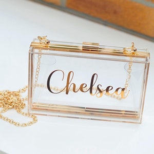 Pode incluir: Uma bolsa de mão transparente de acrílico com uma corrente dourada e um fecho dourado. A bolsa tem o nome "Chelsea" impresso em letras douradas na frente.