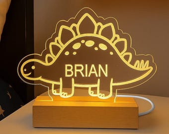 May include: A glowing dinosaur-shaped night light with the name "BRIAN" illuminated in white. The light sits on a wooden base and is plugged in with a white cord. The dinosaur is a light amber color.