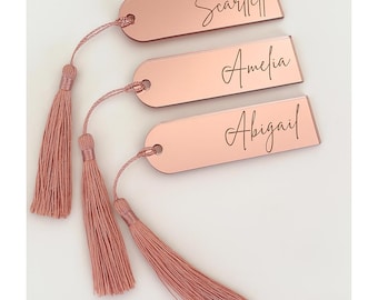 May include: Three rose gold name tags with tassels. Each tag has a different name in a cursive font: Scarlett, Amelia, and Abigail. The tags are rectangular with rounded corners and a small hole for the tassel string.