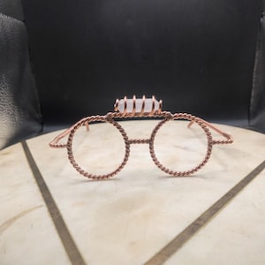 May include: A pair of glasses crafted from twisted copper wire, featuring round frames and a clear, cylindrical quartz crystal secured horizontally across the bridge with copper wire wrapping. The design includes thin, twisted wire temples.