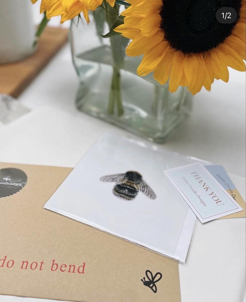 K&ouml;nnte beinhalten: Eine wei&szlig;e Karte mit einer Illustration einer schwarzen und gelben Hummel. Die Karte liegt auf einem braunen Umschlag mit dem Text "Nicht knicken" und einer kleinen Bienenillustration. Eine Dankeskarte mit dem Text "Vielen Dank" liegt auf der Karte.