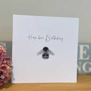 Può includere: Biglietto di auguri quadrato bianco con la scritta "Hap-bee Birthday" in corsivo. Un'illustrazione dettagliata di un'ape è centrata sotto il testo. Il biglietto è su una superficie di legno.