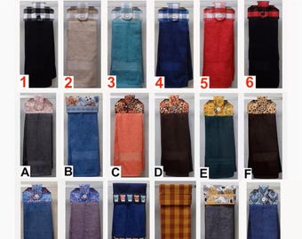 May include: A collection of eighteen hanging kitchen towels labeled 1-6, A-F, and G-M1. Each towel features a fabric topper with a button closure in various patterns, including plaid, floral, and geometric prints, paired with solid or patterned terry cloth.