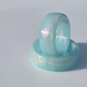 May include: Two light blue resin rings, one stacked on top of the other. The rings have a pearlescent shimmer, catching the light. The rings are a solid band, with a smooth, rounded surface. The rings are a light, pastel blue color.