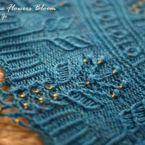 Könnte beinhalten: Nahaufnahme eines blauen gestrickten Schals mit goldenen Perlen. Der Schal hat ein Spitzenmuster und ist mit goldenen Perlen verziert. Der Text "When The Flowers Bloom" ist oben im Bild sichtbar.
