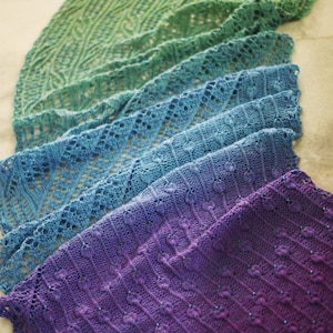May include: A collection of hand-knitted shawls in shades of green, blue, and purple. Each shawl features intricate lace and textured patterns. The shawls are arranged in a layered display, showcasing the gradient of colors and detailed craftsmanship.