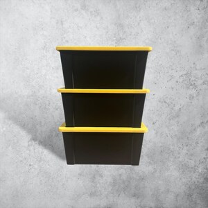 K&ouml;nnte beinhalten: Ein Stapel aus drei rechteckigen Aufbewahrungsboxen, die jeweils einen soliden schwarzen Boden und einen leuchtend gelben Deckel haben. Die Boxen sind vertikal angeordnet und bilden eine einheitliche S&auml;ule mit abwechselnden schwarzen und gelben Streifen.