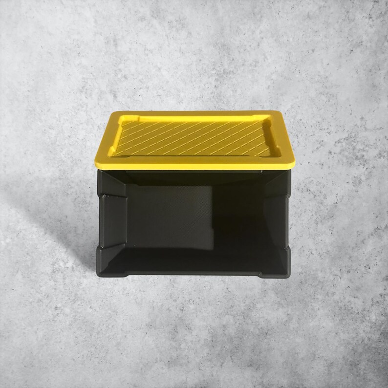 K&ouml;nnte beinhalten: Eine rechteckige Aufbewahrungsbox mit einem soliden schwarzen Unterteil und einem leuchtend gelben Deckel. Der Deckel weist auf seiner Oberfl&auml;che ein erhabenes, diagonal strukturiertes Muster auf. Die Box ist offen und zeigt einen tiefen Innenraum.
