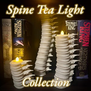 May include: Three white spine-shaped tea light holders with a candle in each. The holders are arranged on a shelf with Stephen King books behind them. The text "Spine Tea Light Collection" is superimposed on the image.
