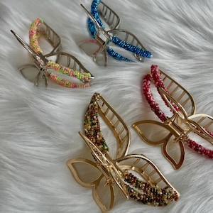May include: A selection of butterfly-shaped hair clips. The clips have a metal butterfly design with beaded detailing in blue, pink, red, green, and brown. They are presented on a white, fluffy background.