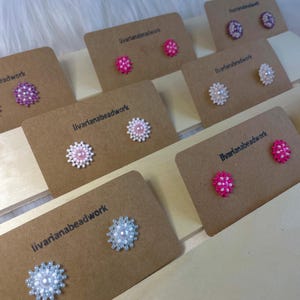 May include: A selection of handmade beaded flower stud earrings in shades of pink, white, and purple. Each pair is presented on a small brown card with the text "livarianebeadwork". The earrings are displayed on a light wooden surface.