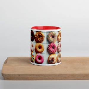 May include: A white ceramic mug with a red rim features a colorful pattern of donuts on a white background. The donuts are arranged in rows and include various toppings, such as sprinkles, chocolate, and frosting.