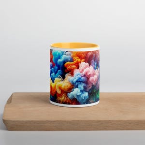 May include: A white ceramic mug with a yellow rim features a colorful abstract design of swirling blue, orange, pink, and yellow paint. The mug is sitting on a wooden surface.