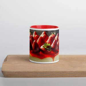 Könnte beinhalten: Eine rote und weiße Keramiktasse mit einem Bild von einem Erdbeer-Käsekuchen. Der Käsekuchen ist mit frischen Erdbeeren und einem roten Guss verziert.