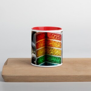 May include: A white ceramic mug with a red rim and a colorful rainbow cake design. The cake is layered with red, orange, yellow, green, and blue frosting.
