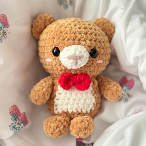 May include: A brown crocheted teddy bear with a white chest and a red bow tie. The bear is lying on a white bedspread with a floral pattern.