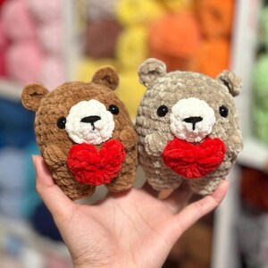 May include: Two handmade crochet teddy bears. One is brown, the other is gray. Both have white faces, black eyes, and red heart-shaped chests. The bears are small and plush, perfect for a gift or decoration.
