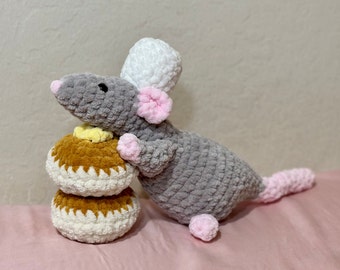 May include: A gray crocheted mouse with pink ears and a white crocheted hat holding two brown and white crocheted donuts.