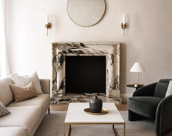Puede incluir: Un salón con chimenea de mármol, espejo redondo dorado y sofá beige. Una mesa de centro blanca con marco dorado sostiene un jarrón oscuro y una bandeja dorada. Un sillón verde oscuro está junto a una pequeña mesa auxiliar con una lámpara.