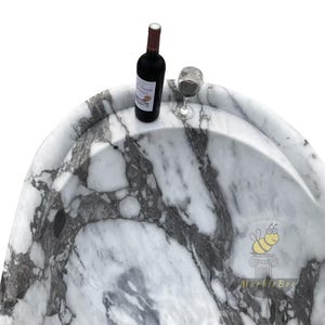 May include: A marble bathtub with a white and gray veined pattern. A bottle of red wine and a wine glass are on the edge of the tub. The tub has a curved design and a smooth surface.