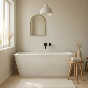 May include: A minimalist bathroom scene featuring a white marble bathtub, a wooden stool with towels and candles, and a beige rug. A wooden-framed mirror hangs above the tub, and a white pendant light hangs from the ceiling. The walls are a neutral beige.