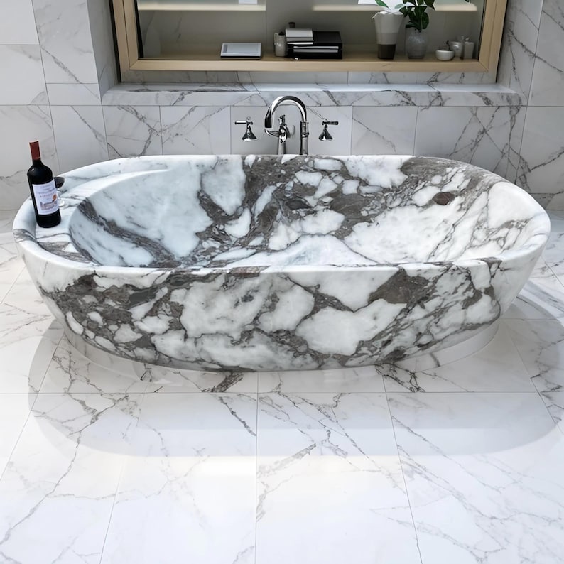 May include: Oval-shaped marble bathtub with gray veining. The tub sits on a white marble floor. A bottle of red wine rests on the tub's edge. The bathroom features a large mirror and a chrome faucet.