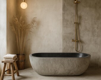 May include: A modern bathroom with a stone-like oval bathtub with a dark interior. A wooden stool holds folded towels. A gold shower fixture is mounted on a concrete wall. Decorative pampas grass in a wooden pot adds a natural touch.
