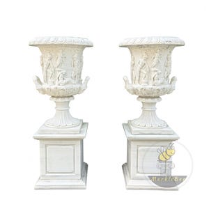 May include: A pair of white marble urns, each with classical relief carvings and a square pedestal base. The urns feature a wide, fluted rim and decorative handles. The design is inspired by ancient Greek or Roman aesthetics.