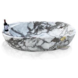 May include: Oval-shaped marble bathtub with a white and gray veined pattern. A small bottle of red wine sits on the edge of the tub. The tub has a built-in shelf and a smooth, polished finish.