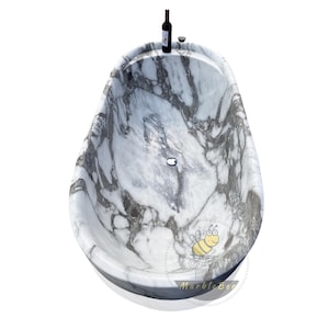 May include: A marble bathtub with a gray and white pattern, oval-shaped, and a built-in drain. A bottle of red wine and a wine glass are placed on the rim. The tub has a glossy finish and is set against a white background.