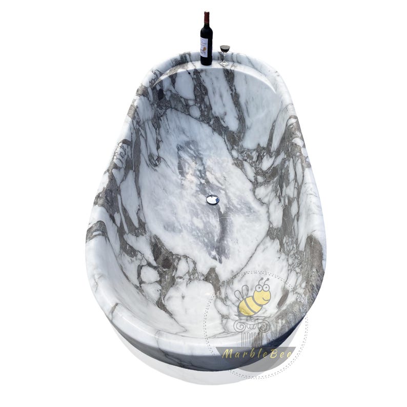 May include: A marble bathtub with a gray and white pattern, oval-shaped, and a built-in drain. A bottle of red wine and a wine glass are placed on the rim. The tub has a glossy finish and is set against a white background.