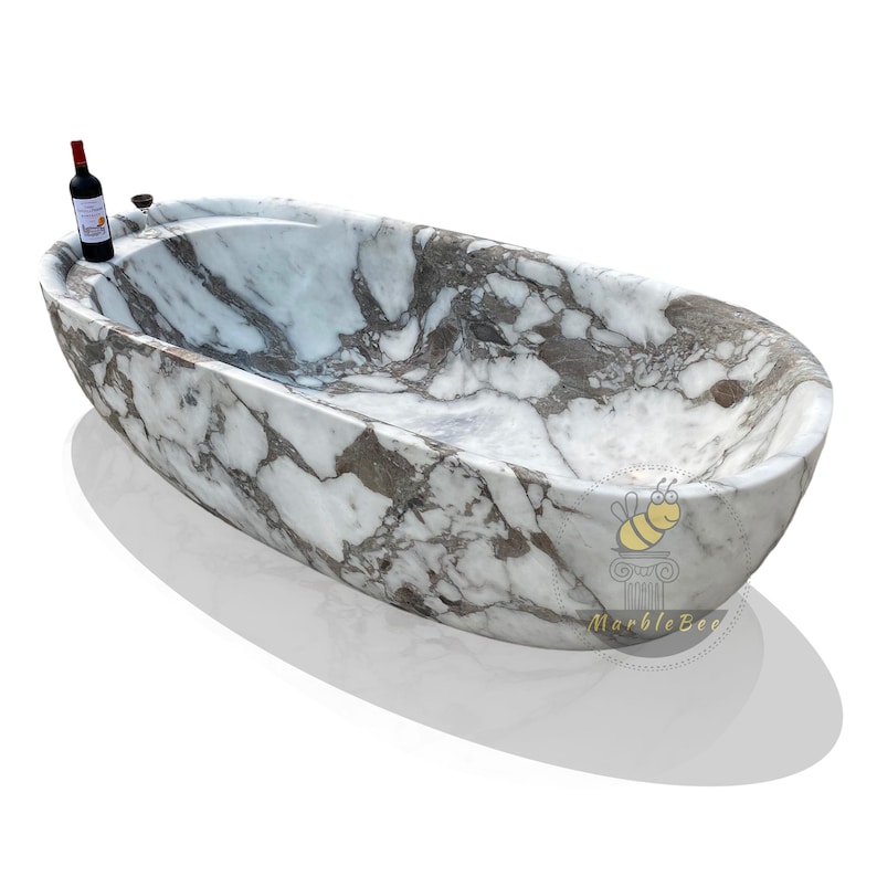 May include: Oval-shaped marble bathtub with a white and gray veined pattern. A bottle of red wine and a glass are placed on the rim. The tub has a smooth, polished finish and is set against a white background.