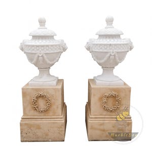 May include: A pair of white marble urns with intricate carvings, featuring lion heads and garland patterns. Each urn rests on a beige square pedestal, adorned with a carved wreath. The urns have a classic, elegant design.