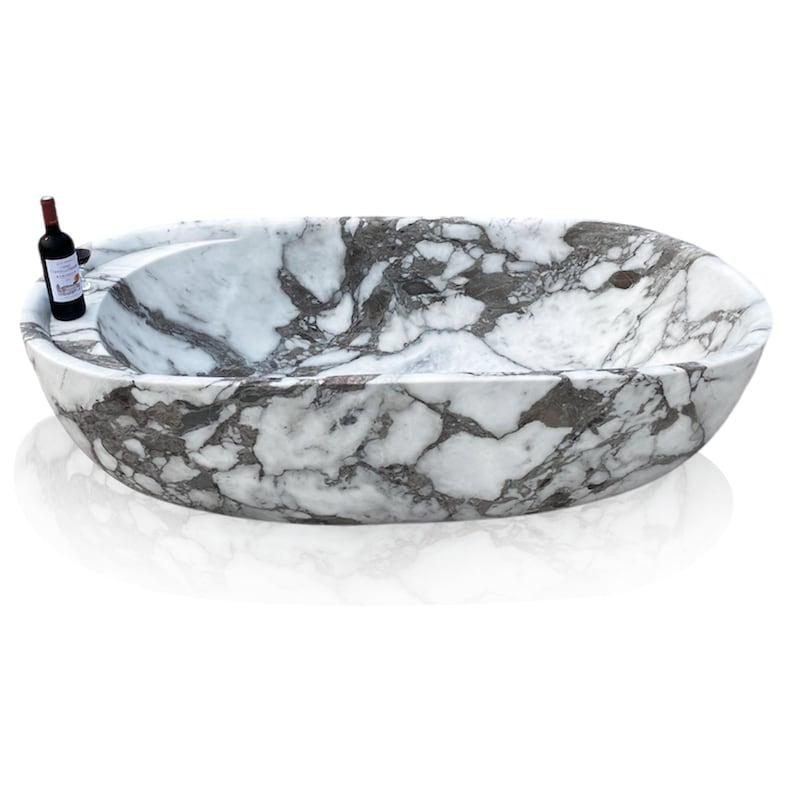 May include: Oval-shaped marble bathtub with a white and gray veined pattern. A small bottle of red wine rests on the edge of the tub. The tub has a smooth, polished finish and a built-in ledge.