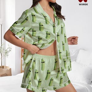 Könnte beinhalten: Hellgrünes Pyjama-Set mit einem sich wiederholenden Muster von Cartoon-Gurken mit Sonnenbrille. Das Set besteht aus einem kurzärmeligen Hemd mit Knöpfen und passenden Shorts, beide mit weißem Paspel versehen. Das WXRIDE-Logo ist sichtbar.