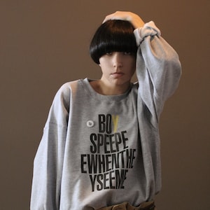 May include: A gray sweatshirt with the text "BOY SPEEPE EWHENTHE YSEEME" in black and yellow. The person is wearing khaki pants and a brown belt. A small pin is attached to the sweatshirt.