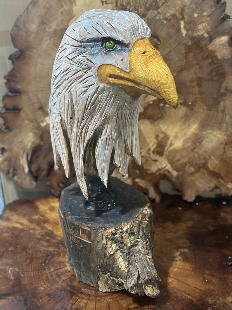 May include: A hand-carved wooden sculpture of a bald eagle's head with a detailed, realistic design. The eagle's feathers are carved with intricate detail, and its eyes are painted a bright green. The sculpture is mounted on a wooden base.