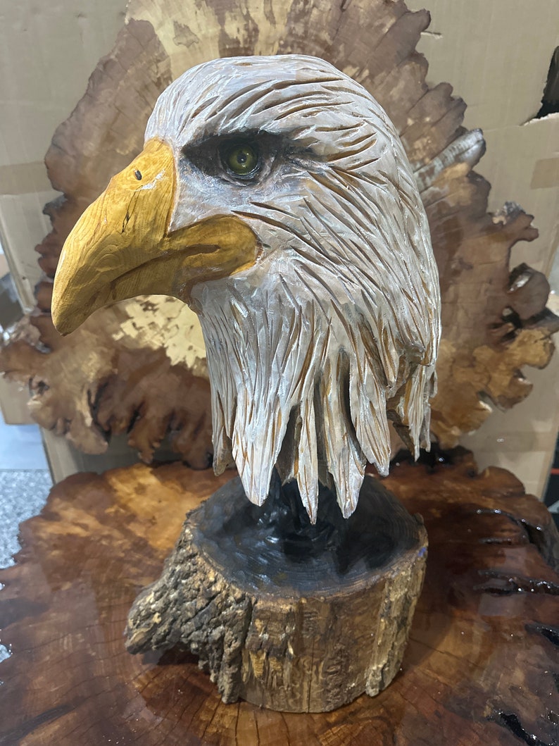 May include: A hand-carved wooden sculpture of a bald eagle's head, with realistic details and a natural wood finish. The eagle's head is mounted on a wooden base.