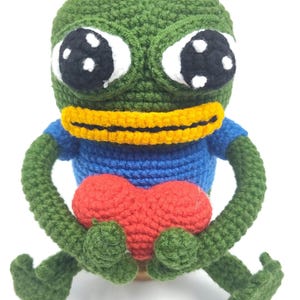 May include: A handmade crochet frog doll in green, blue, yellow, white, and red. The frog has large white eyes with black pupils, a yellow collar, and a blue shirt. It is holding a red heart and has green arms and legs.