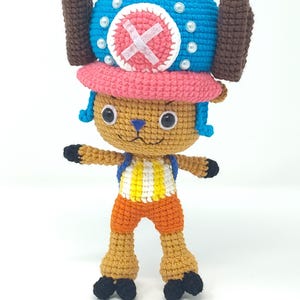 May include: A handmade crochet figure of a cartoon character. The figure has a blue hat with white accents, a pink brim, and brown antlers. It wears an orange outfit with a yellow and white striped shirt. The figure has black feet and hands.