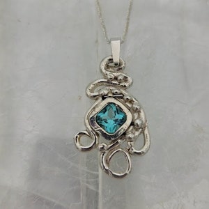 Puede incluir: Un collar con un colgante de plata con una piedra preciosa azul engastada en forma cuadrada. El colgante est&aacute; hecho de alambre de plata retorcido y tiene una cadena delicada.
