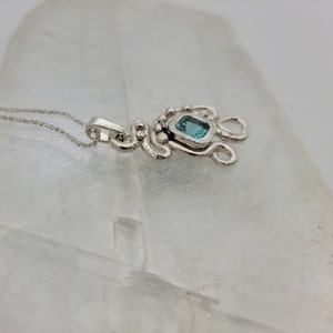 Puede incluir: Un collar de plata con un colgante que tiene una piedra preciosa azul engastada en un marco de plata. El colgante est&aacute; unido a una cadena de plata.