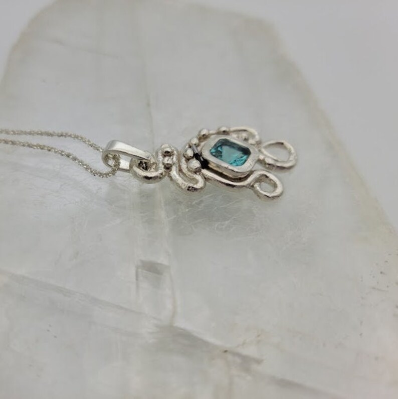 Puede incluir: Un collar de plata con un colgante que tiene una piedra preciosa azul engastada en un marco de plata. El colgante est&aacute; unido a una cadena de plata.