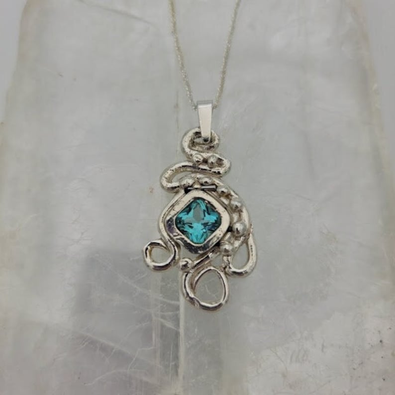 Puede incluir: Un collar con un colgante de plata con una piedra preciosa azul engastada en forma cuadrada. El colgante est&aacute; hecho de alambre de plata retorcido y tiene una cadena delicada.