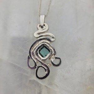 Puede incluir: Un collar con un colgante de plata con una piedra preciosa azul engastada en una forma cuadrada. El colgante est&aacute; hecho de un dise&ntilde;o abstracto y en espiral.