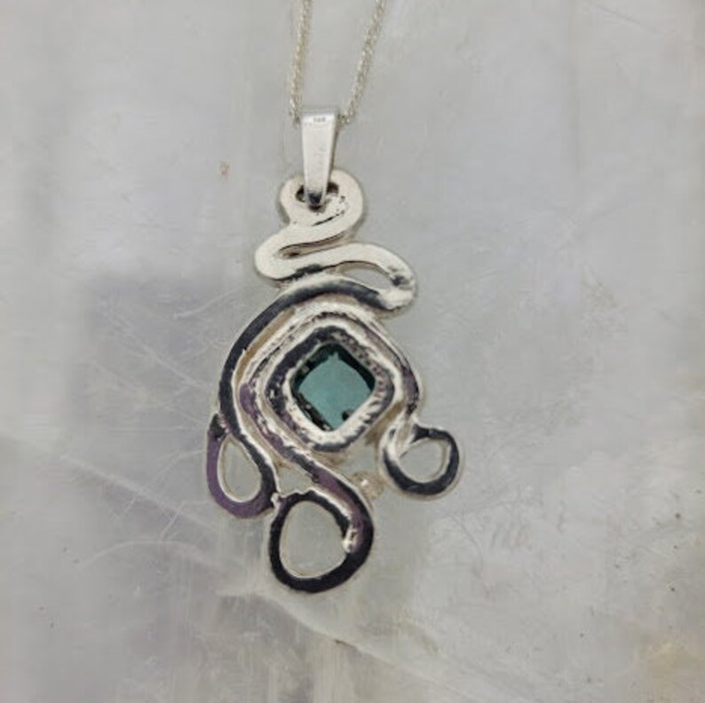 Puede incluir: Un collar con un colgante de plata con una piedra preciosa azul engastada en una forma cuadrada. El colgante est&aacute; hecho de un dise&ntilde;o abstracto y en espiral.